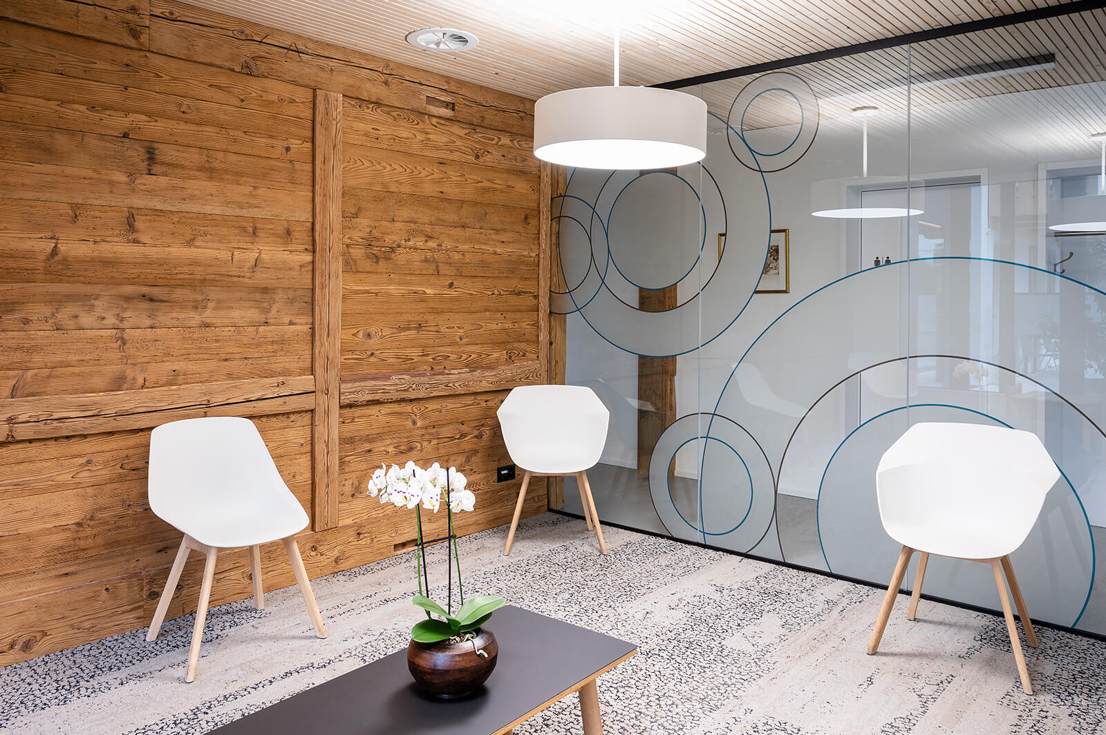 Businessfotografie für die 3A Schreinerei: Moderner Warteraum mit Holzwand und Glasfront Heller Warteraum mit rustikaler Holzwand, Glasfront mit Kreismotiven, drei weissen Stühlen und Tisch mit Orchidee